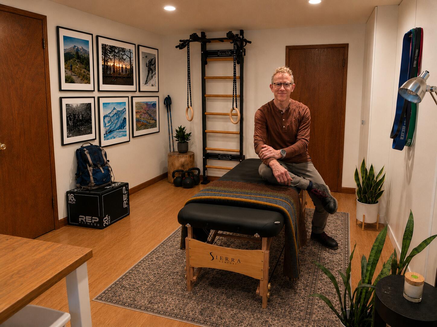 Brian sitting on the treatment table in the wildPaTh studio — blanket, wall bars, kettlebells, and a gallery of adventure photos
